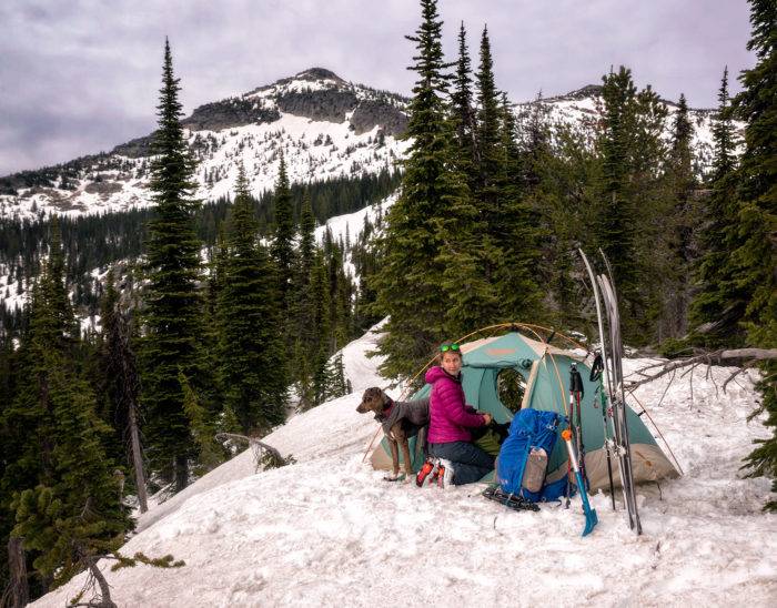 A woman and a dog sitting next to a tent in the snow on top of a mountain.