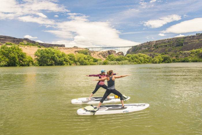Two women practicing yoga on stand-up paddleboards on smooth river with blue skies, canyon walls and an arched span bridge in the distance.