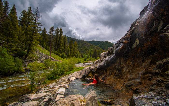 woman sitting in a mountainside hot spring facing a river and tree-covered mountains