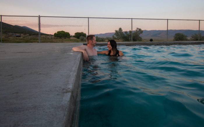 man and woman in an outdoor pool lined by a metal fence and trees and mountains in the background