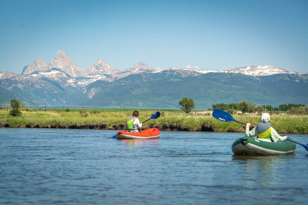 two people in inflatable kayaks paddling in a river with Teton Mountains in the background