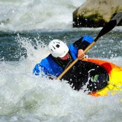 Rapids splash, water droplets cascading around a bright kayak as a man wearing a helmet leans to the side, holding his paddle in his hand.