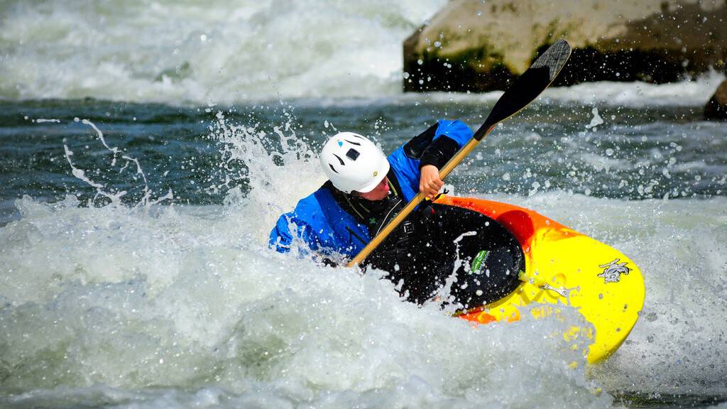 Rapids splash, water droplets cascading around a bright kayak as a man wearing a helmet leans to the side, holding his paddle in his hand.