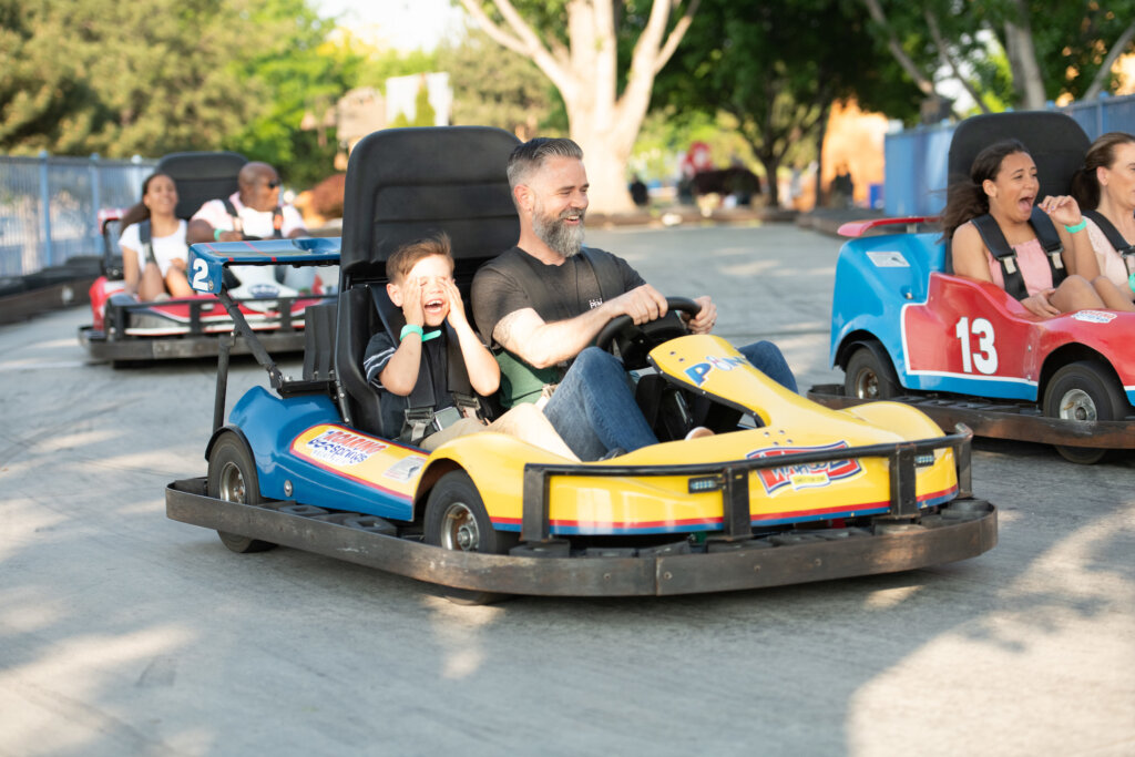 Father and son, both wearing black t-shirts ride in a blue and yellow Go-Kart on a wide track with two other Go-Karts; one to the left and another behind. The dad is smiling and the boy has his hands over his eyes with a laughing smile.