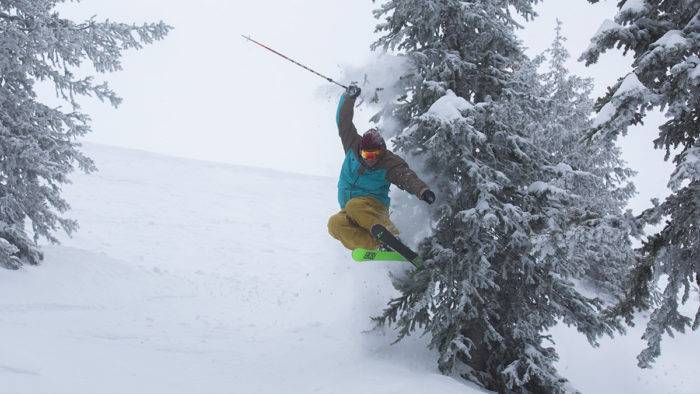 Skier flies over a bump, brushing a snow-covered pine tree causing a poof of powder.