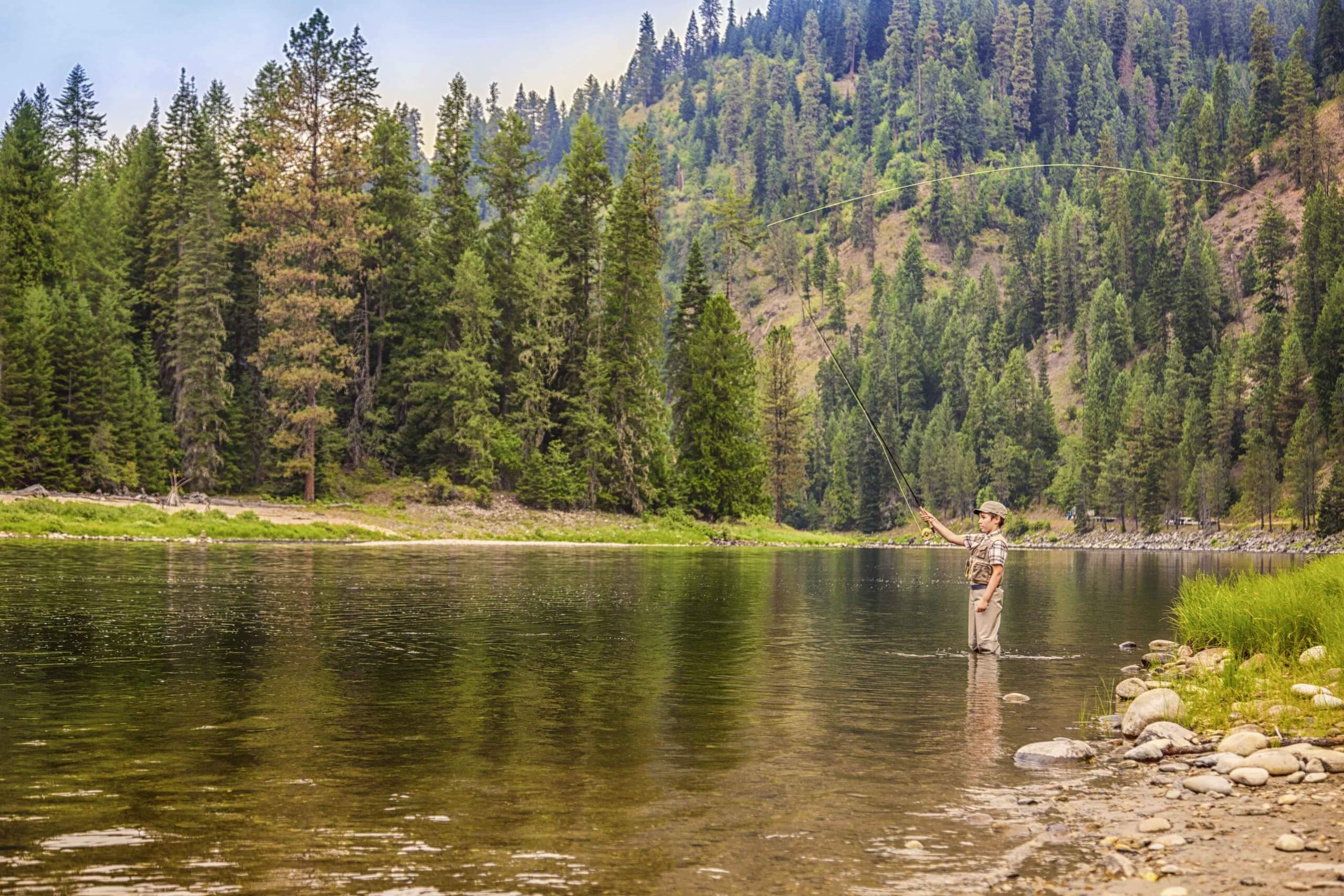 A boy casts a fly fishing line in a calm river.