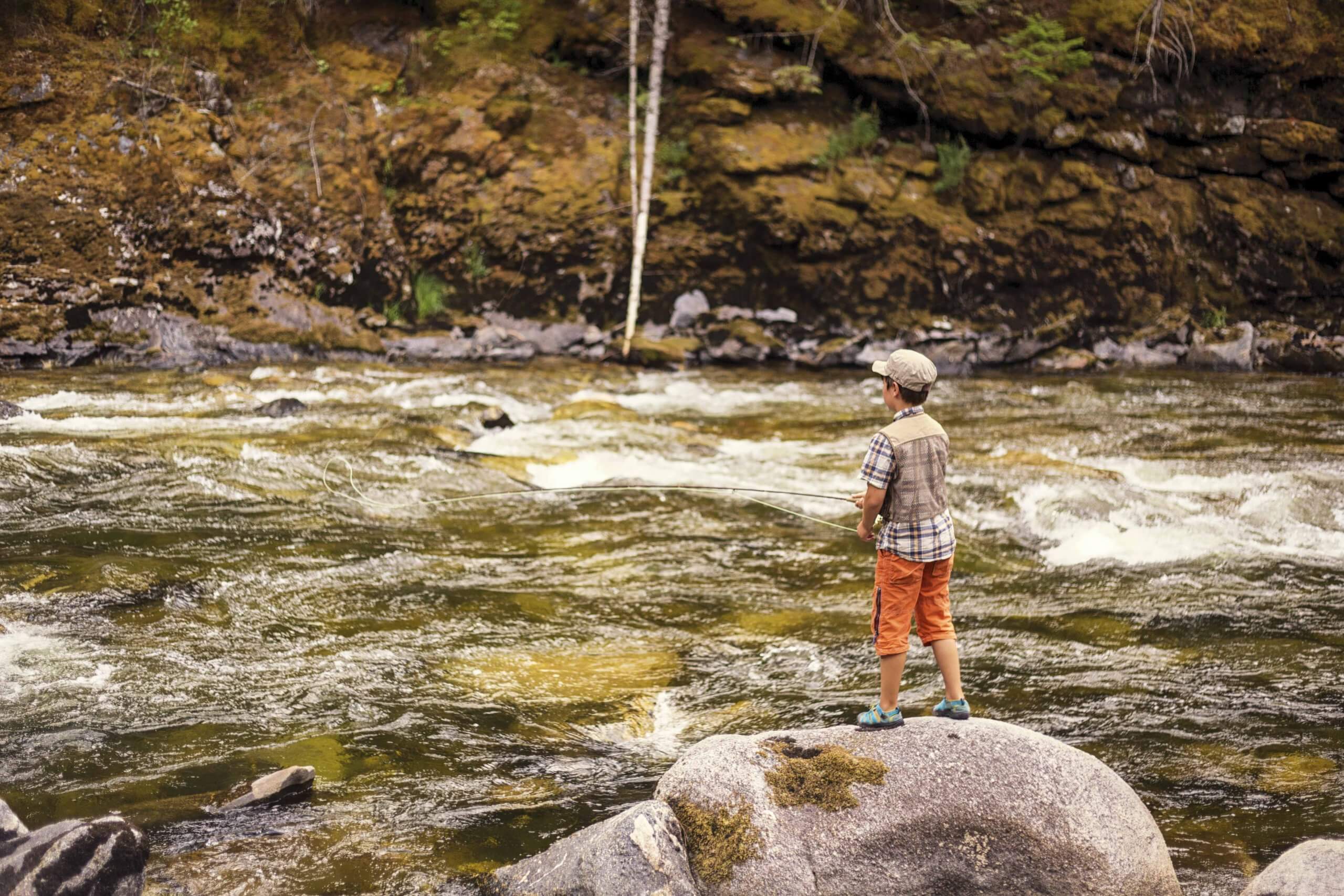 A boy fishing rapids from a rock.