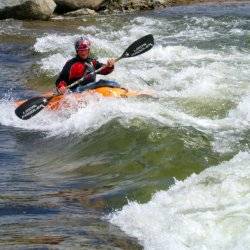 A man holds an oar on an orange kayak as the waters rush around him in Kelly's Whitewater Park, rocks standing behind him.