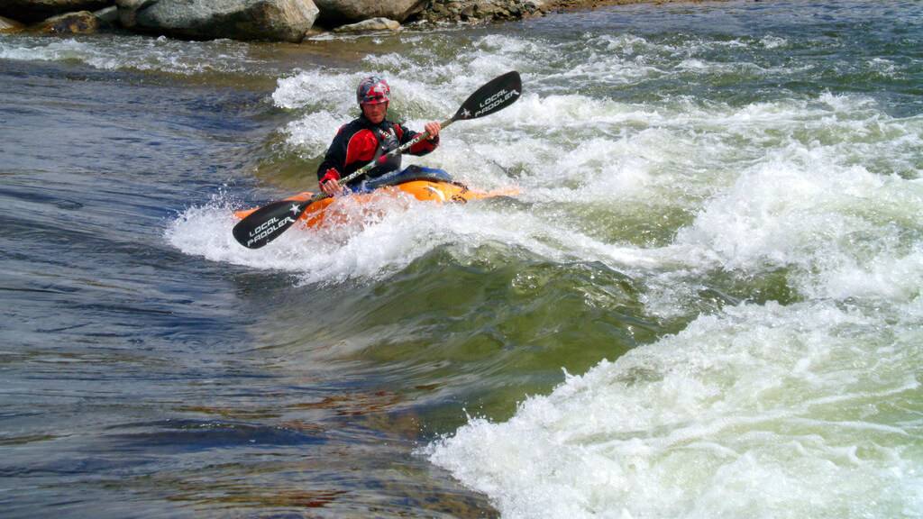 A man holds an oar on an orange kayak as the waters rush around him in Kelly's Whitewater Park, rocks standing behind him.