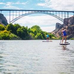 A stand up paddle boarder and a kayaker paddling beneath a large bridge.