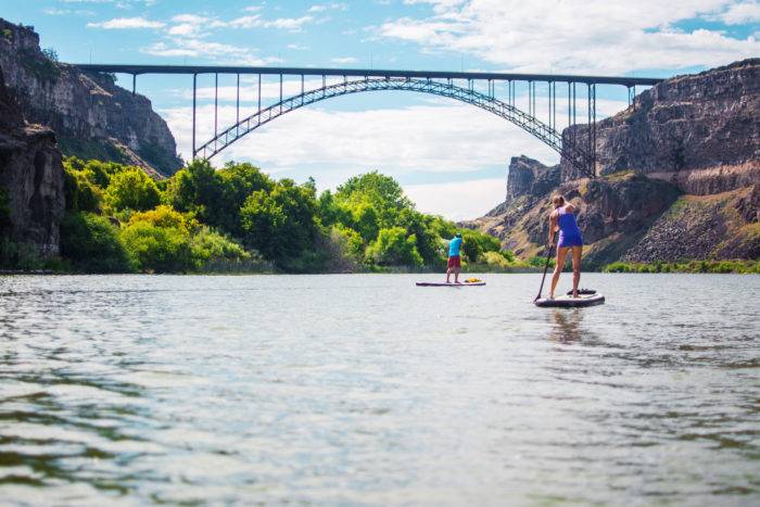 A stand up paddle boarder and a kayaker paddling beneath a large bridge.