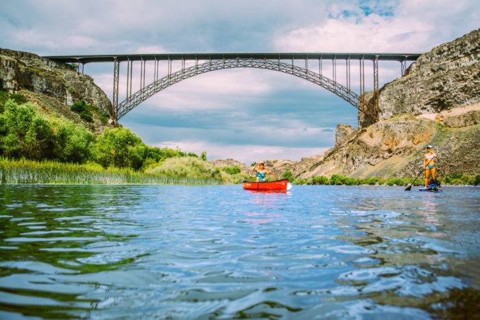 A stand up paddle boarder and a kayaker paddling beneath a large bridge.