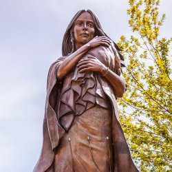 A bronze statue of Sacajawea holding a baby, with trees in the background.