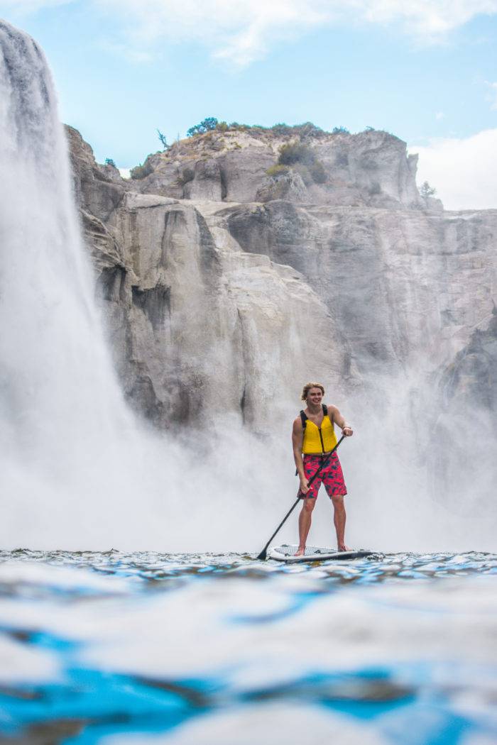 A man SUP'ing beneath a waterfall.