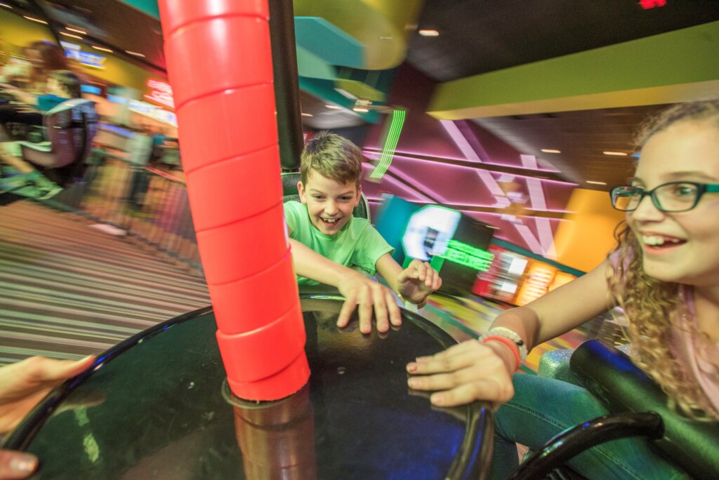 This photo shows movement of a boy, wearing a green shirt, and a girl, wearing pink, on the Twister ride. Both are smiling. The background is slightly fuzzy do to movement with bright colors of pink, yellow and teal.