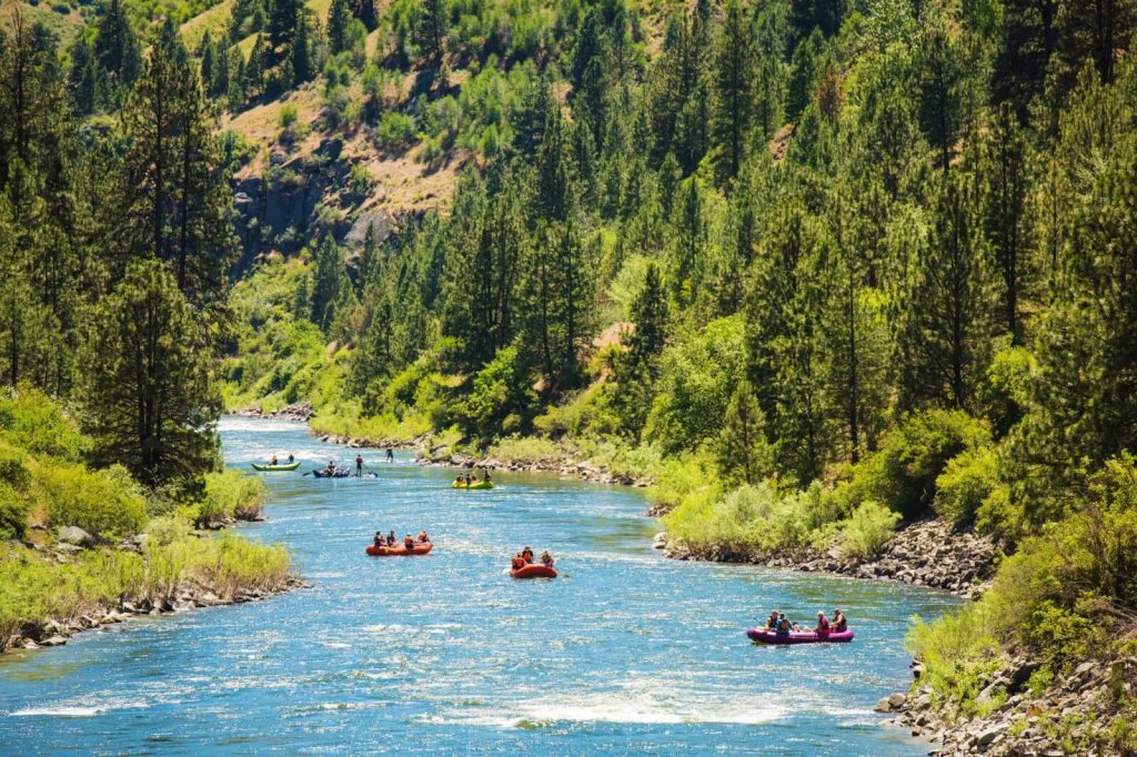 A blue river, dotted with orange and yellow rafts flows between forested, mountainous terrain.