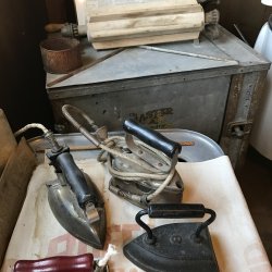 museum display with three metal irons for clothing and a vintage mental wash basin.