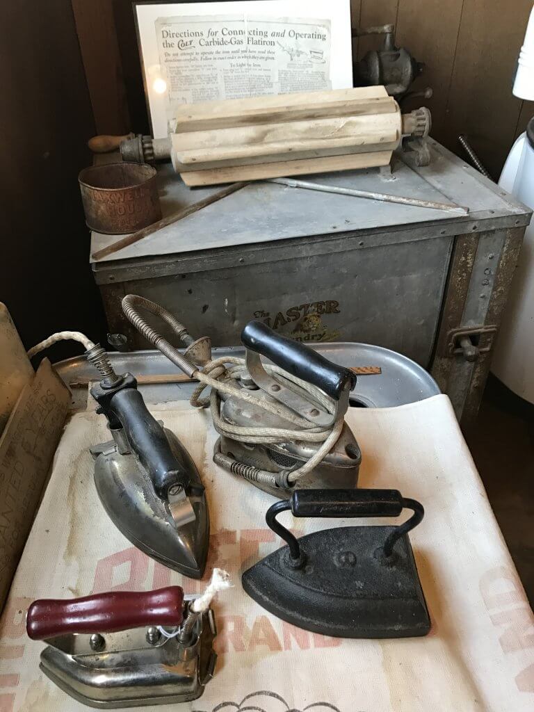 museum display with three metal irons for clothing and a vintage mental wash basin.