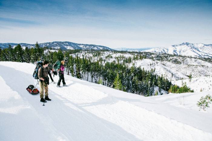 Two people snowshoeing to a yurt along a mountain trail and a forest of trees in the background.