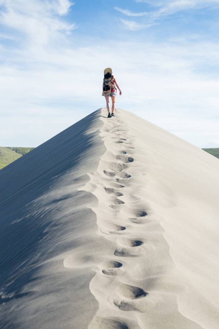 Woman hiking sand dune with sand board on her back.