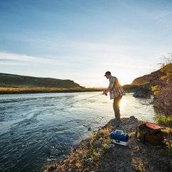 A man fishing along a river at sunset.