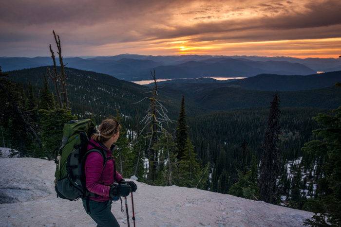 A hiker watches the sun set from the top of a snow covered mountain.