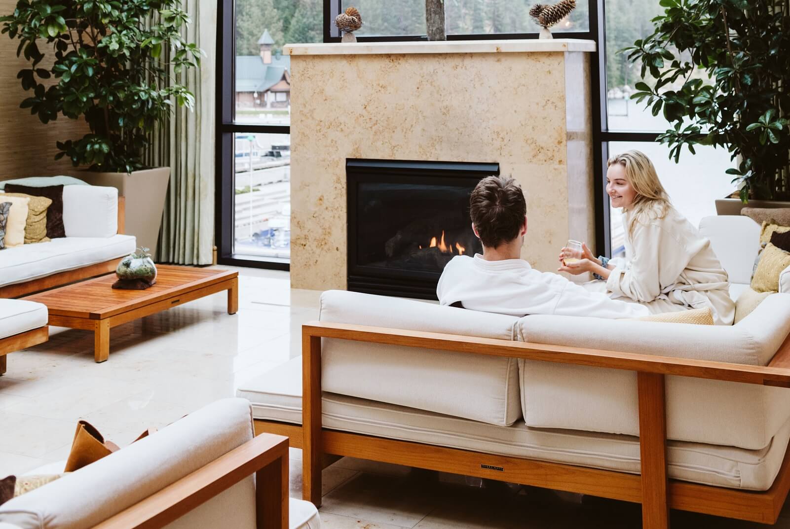 A man and woman relax in plush white spa robes in a room with floor to ceiling windows and a fireplace.