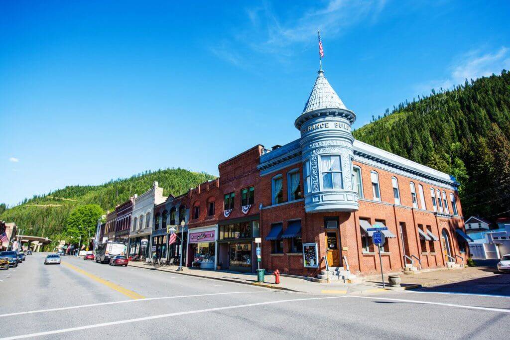 A close-up of the Silver Insurance Company building in Wallace, Idaho, with nearby colorful shops, passing cars, and green mountains behind.