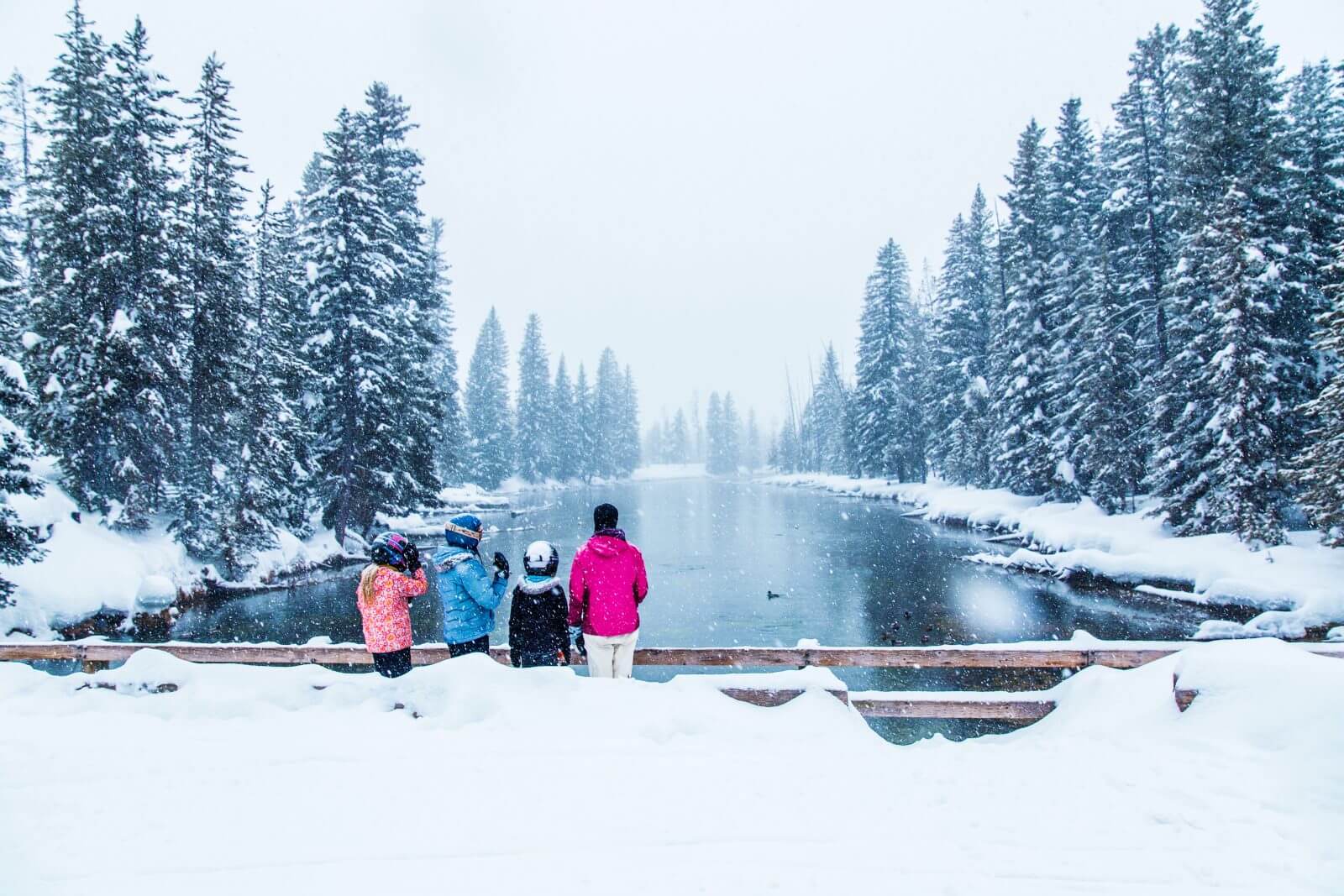 A family of four wearing colorful winter jackets stand on a snow-covered bridge looking at a panoramic view of a river with snow-covered trees lining the river's edge.