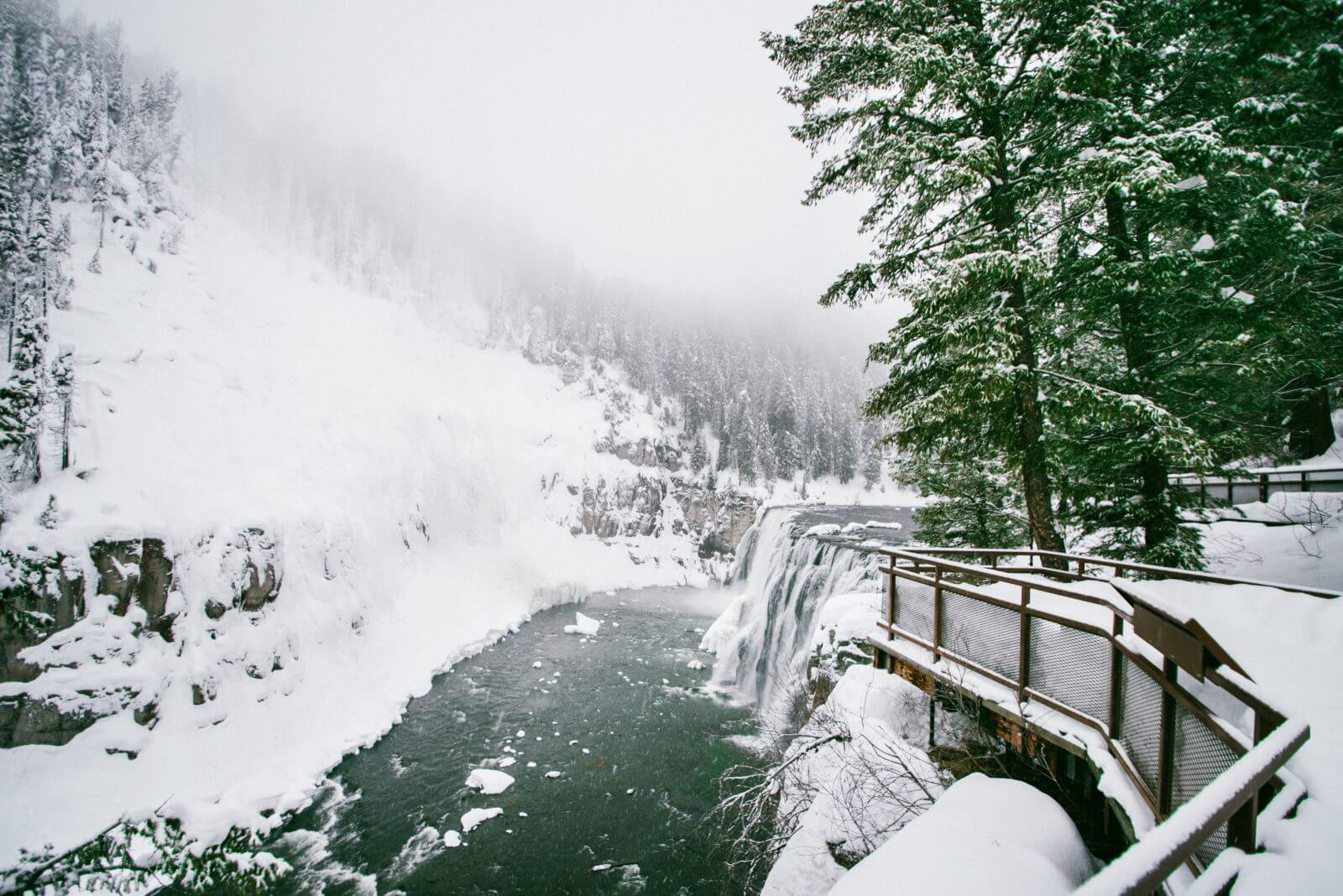 A waterfall cloaked in snow, with a dark green river in the forefront and misty skies above.