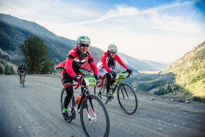 Two bikers ride along a gravel road surrounded by views of mountains and a valley below.