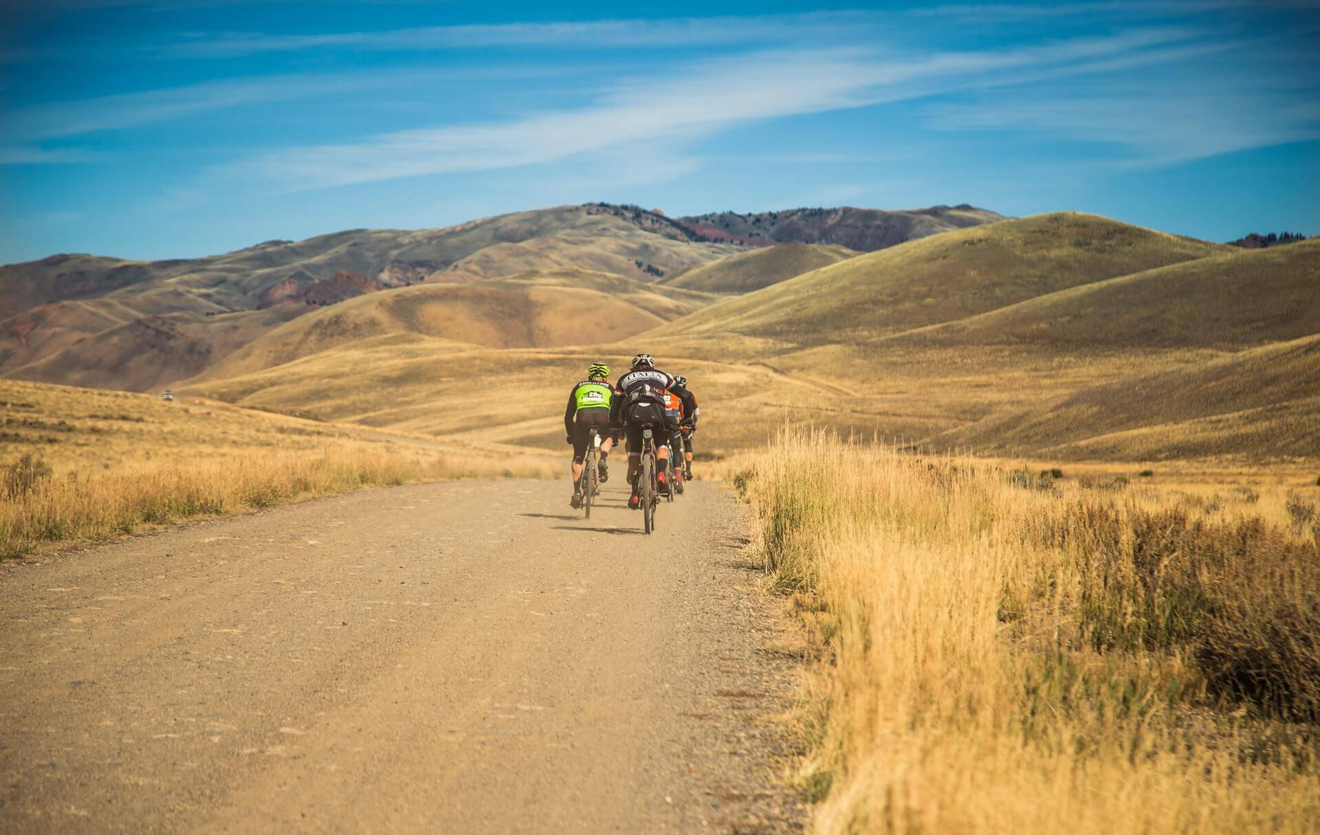 Small group of riders ride on a gravel road surrounded by amber grasses, with mountains and blue skies ahead.