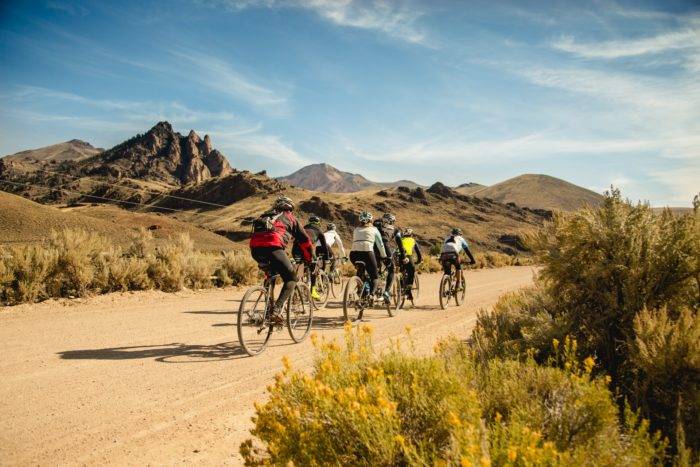 Rider group pedals along dirt road with mountains in the background.