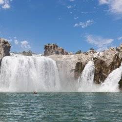 Kayaker raises oars at a far distance in front of the cascading Shoshone Falls under a blue sky.