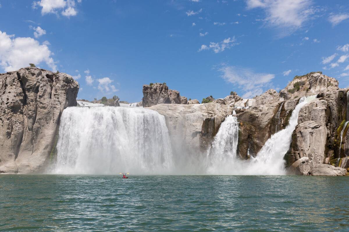 Kayaker raises oars at a far distance in front of the cascading Shoshone Falls under a blue sky.