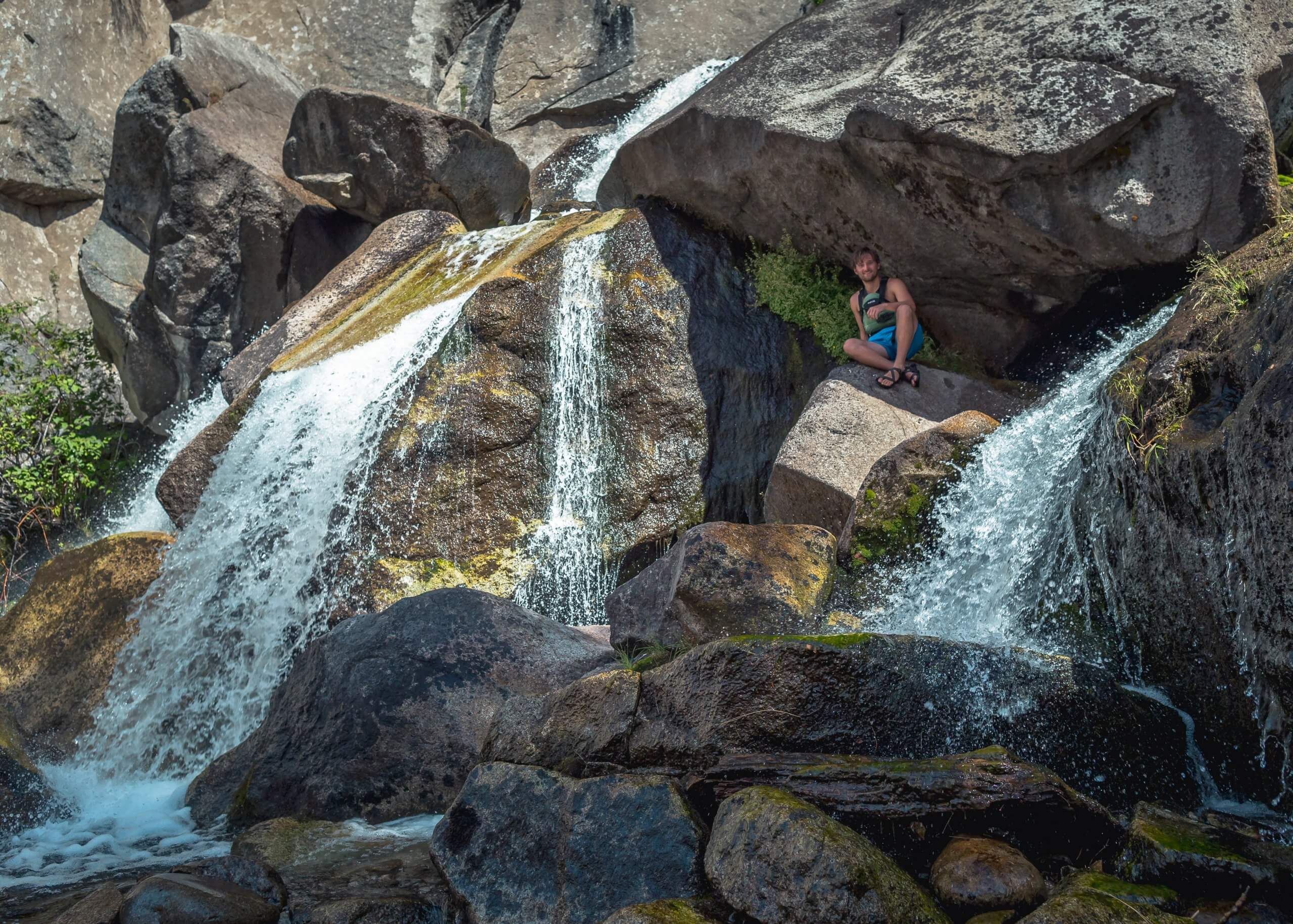 A person sitting on a rock beside a group of large rocks with waterfalls spilling over them at Waterfall Creek.