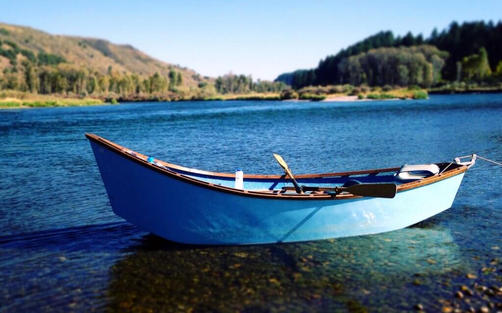 A blue drift boat sits atop the bright waters of the South Fork of the Snake River where green trees and mountains tower in the distance.