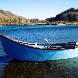 A blue drift boat sits atop the bright waters of the South Fork of the Snake River where green trees and mountains tower in the distance.