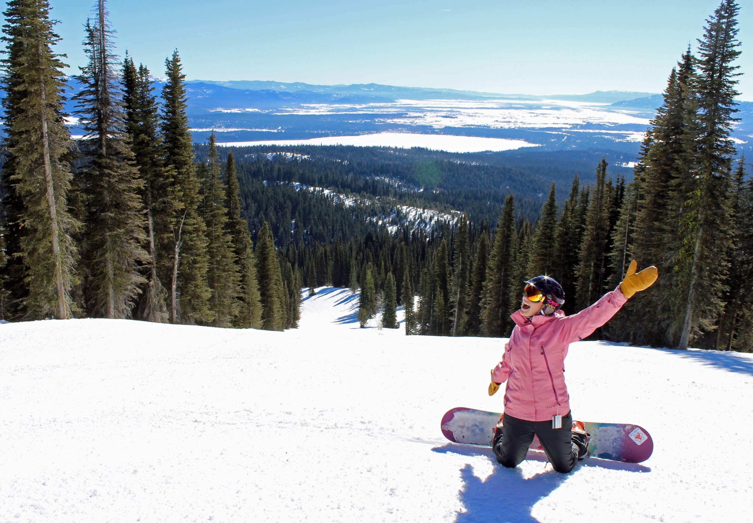 A woman in pink kneels and smiles at the top of a slope at Brundage Mountain Resort.
