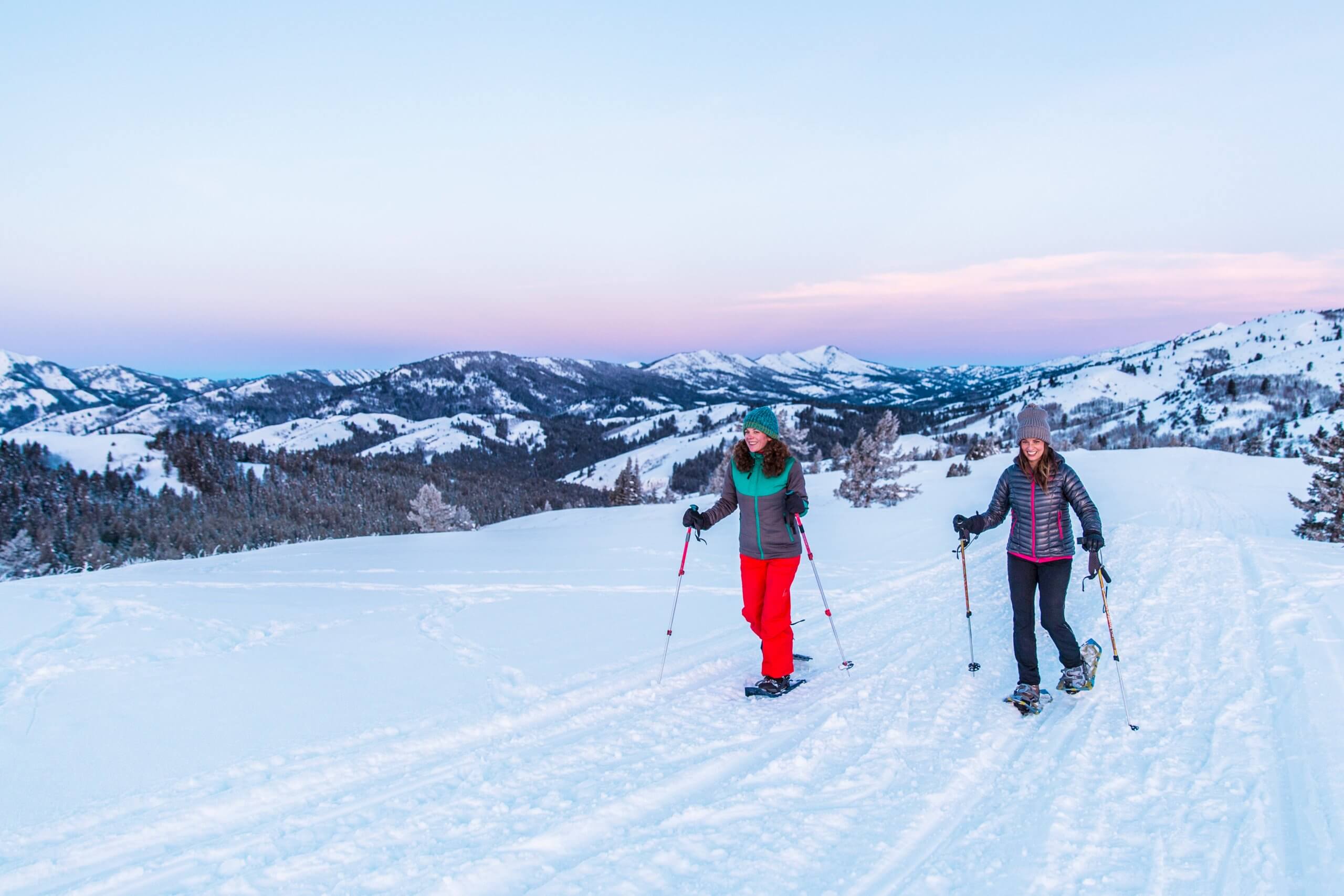 Two women in puffer jackets and beanies snowshoe with snow-covered mountains in the background in Victor, Idaho.