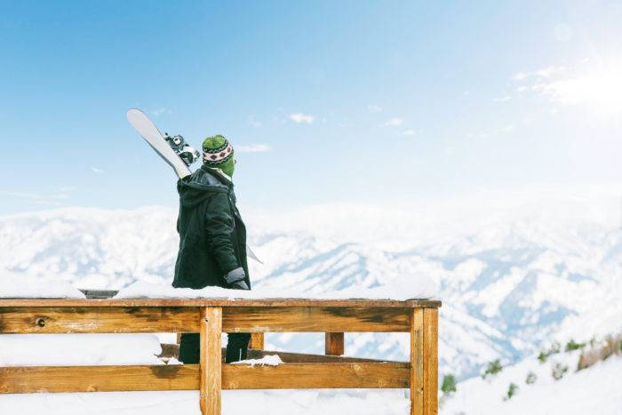 A snowboarder standing on the deck of a yurt overlooking a snow-covered valley.