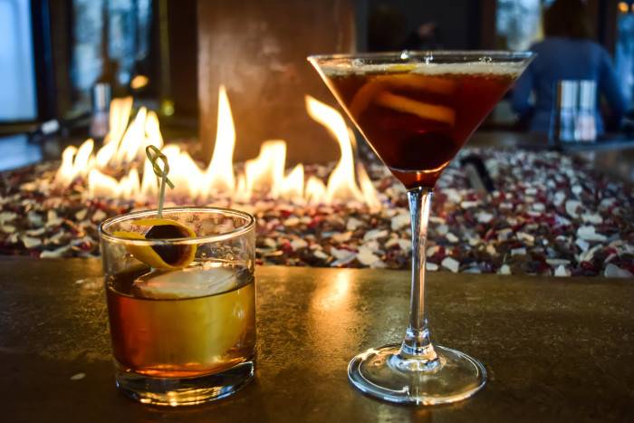 Two cocktails, one a light amber in a hi-ball glass and the second a dark amber with orange peel visible in a martini glass, reflect the flickering from a firepit at 13th Street Pub in Boise.