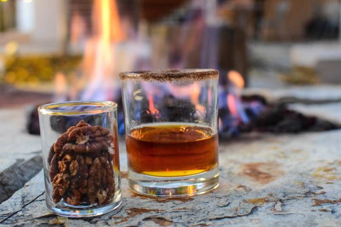 A dark amber cocktail half fills the glass rimmed with a nutty colored salt and sits next to a small glass of spiced pecans with a glowing fire from the fire pit in the background at the Modern Hotel in Boise.