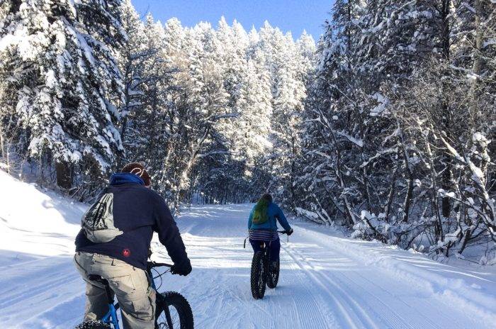 Man and woman biking on the nordic trails at Kelly Canyon, surrounded by snow-covered trees and a bright blue sky.