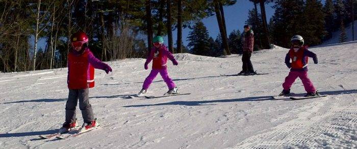 Young children bright colored ski school vests ski down an easy slope.