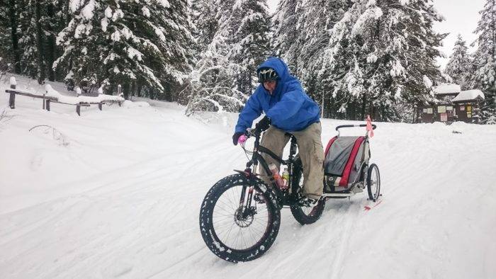 Man pedals fat bike pulling a bike trailer on sleds behind it.