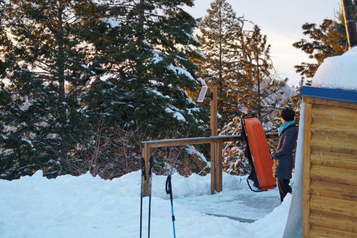 Woman gazes from the deck of the yurt and the surround snow and scenery, with cross country ski poles in the foreground.