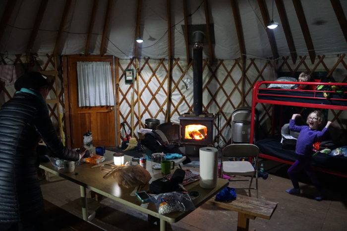 Mom working on dinner with kids and a warming fire in the wood stove in the foreground.