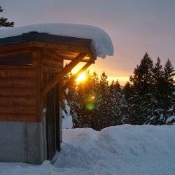 Snow covers the roof and overhang of the yurt's shed while the sun sets in the background.