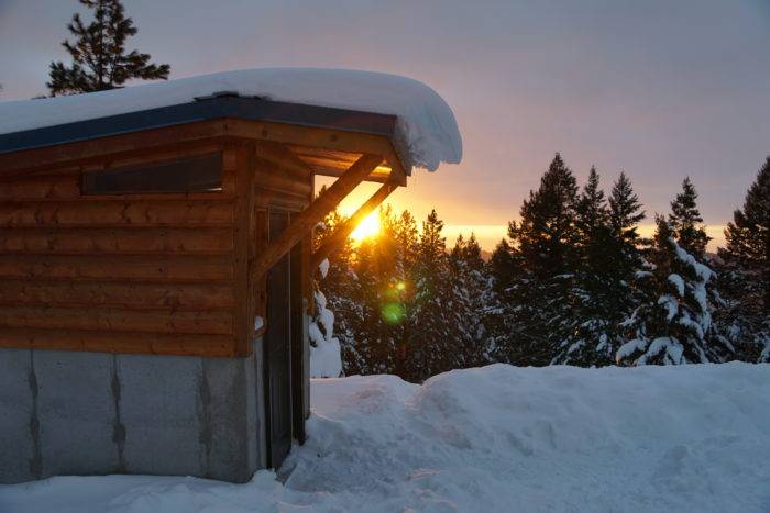 Snow covers the roof and overhang of the yurt's shed while the sun sets in the background.